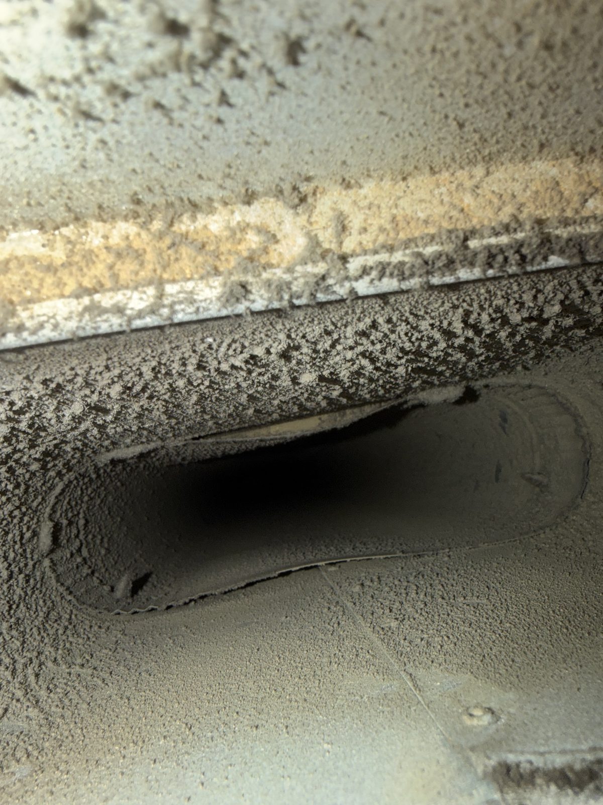 Thick gray dust coating the inside of a duct around an oval takeoff, before cleaning