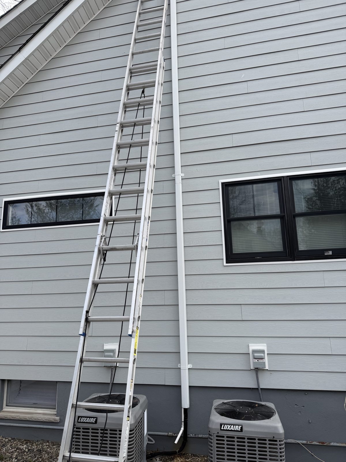 An extension ladder raised against gray siding beside two air conditioning condensers