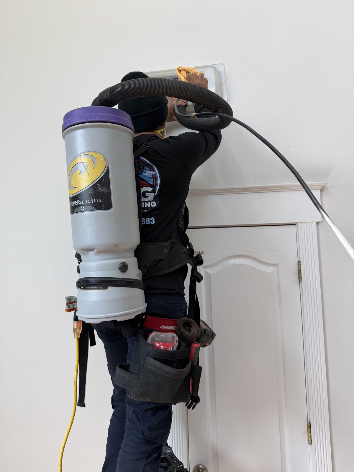 An AMG technician wearing a backpack vacuum, cleaning a wall vent above a doorway