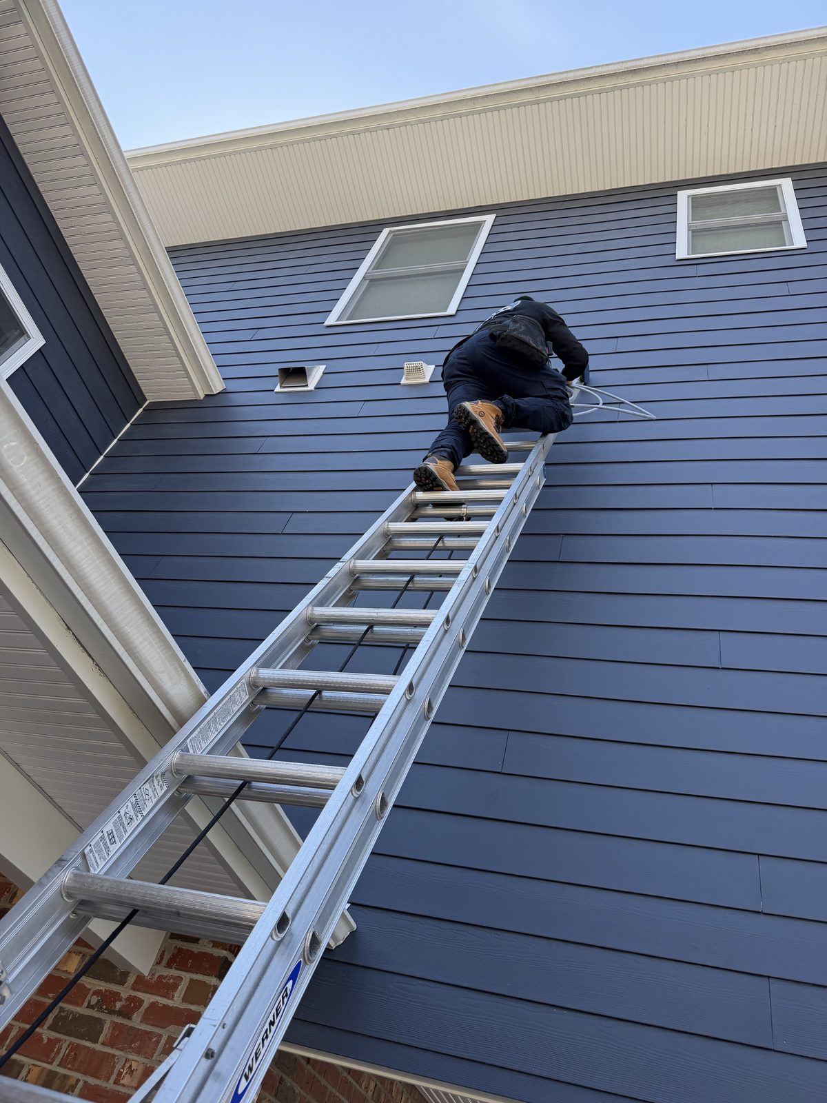 An AMG technician high on an extension ladder, reaching a vent on blue siding