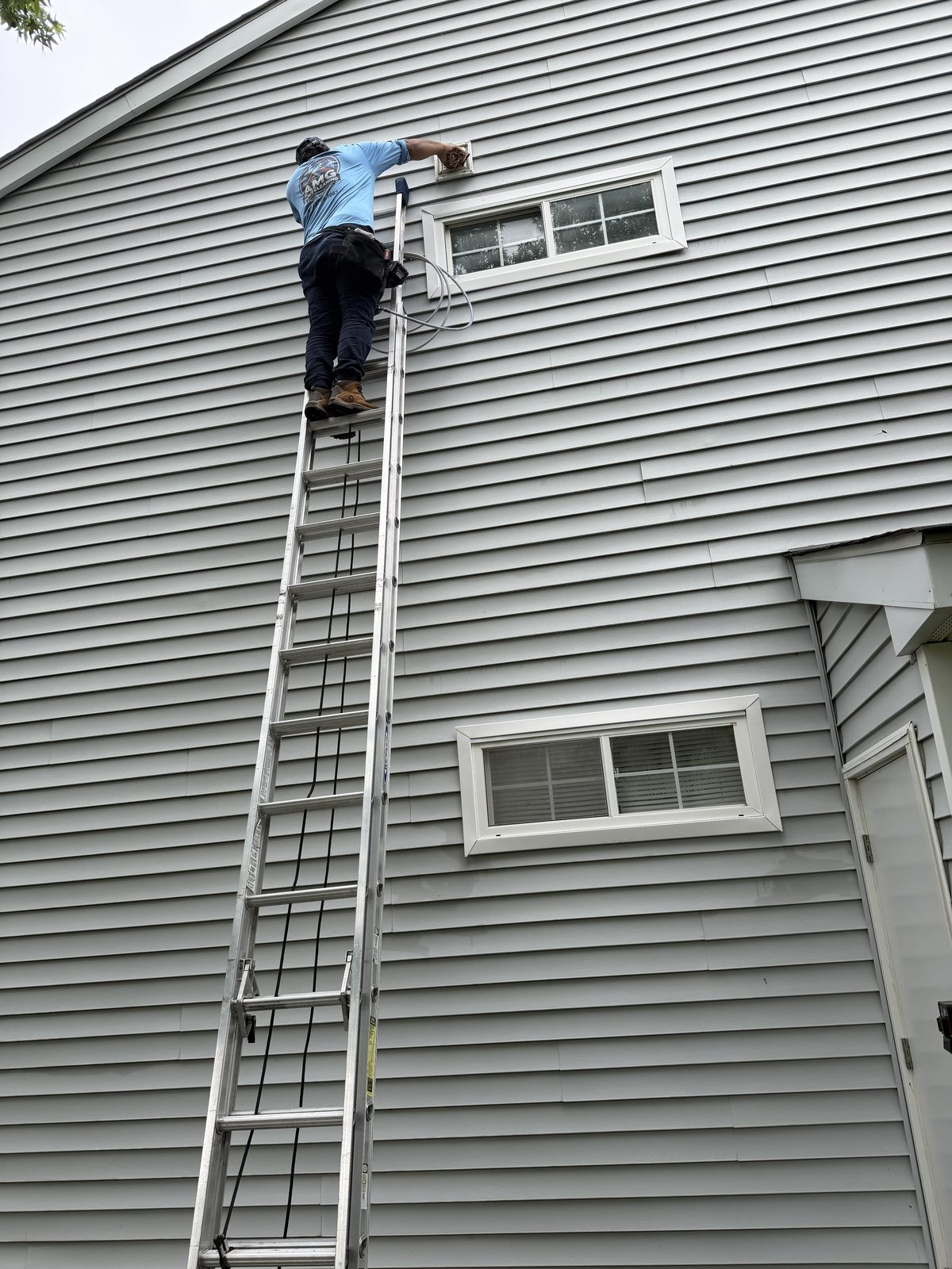 An AMG technician at the top of a tall extension ladder, working at a vent on gray siding