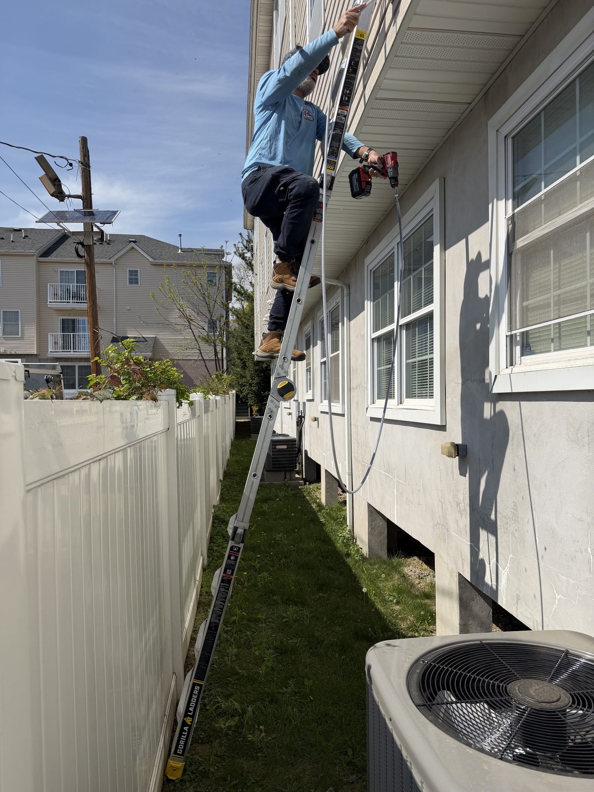 An AMG technician on a ladder beside a row of townhouses, drill in hand at an upper vent