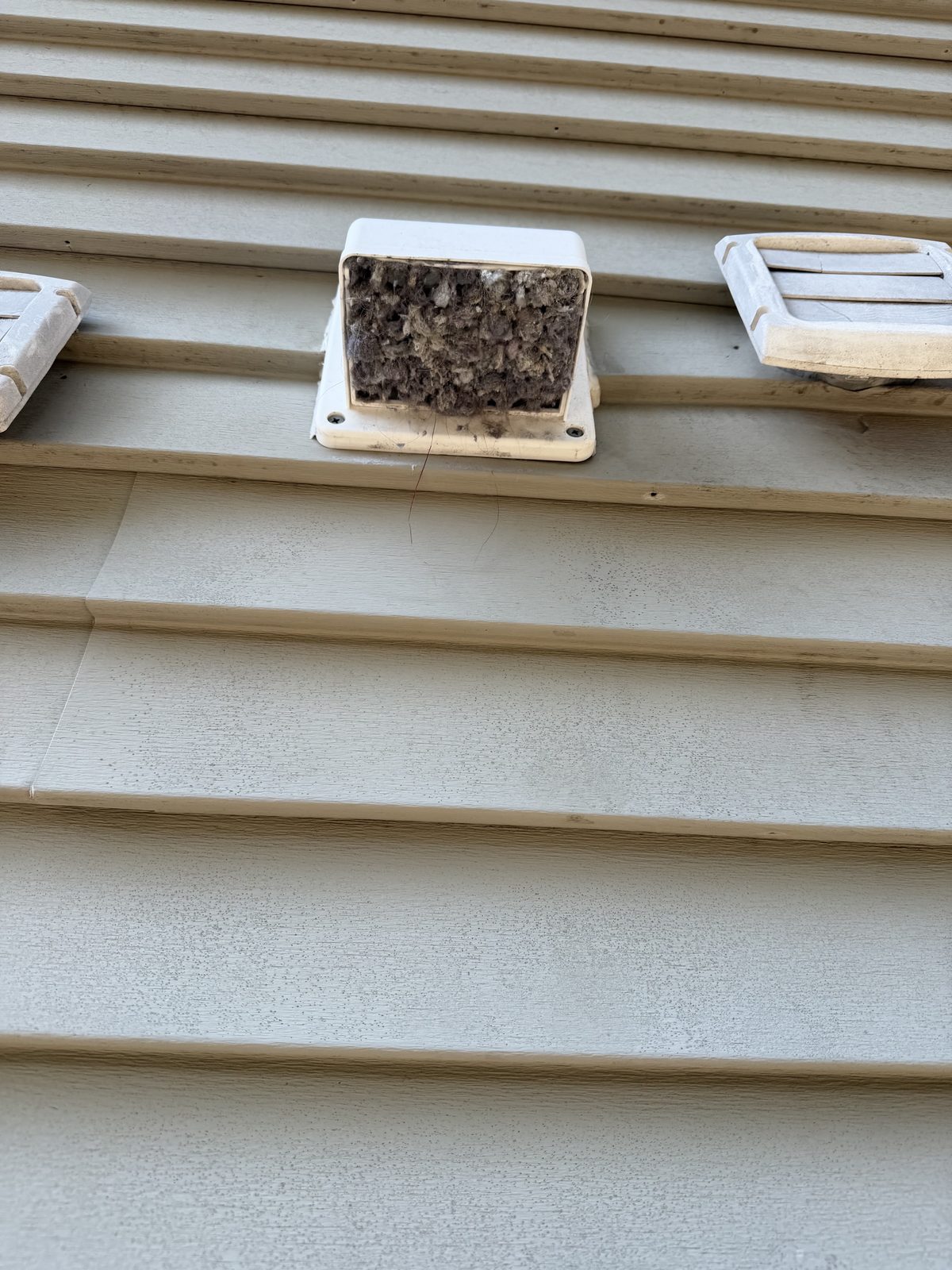 Three vent hoods on beige siding, the middle one packed solid with lint
