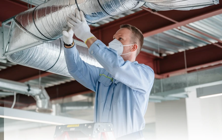 HVAC technician in a light blue uniform and mask working on a metallic air duct from a ladder in an industrial-style space